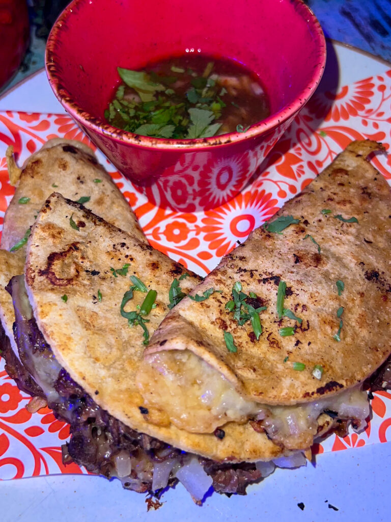 Birria tacos with a consume in a red pot on a plate from The Basement